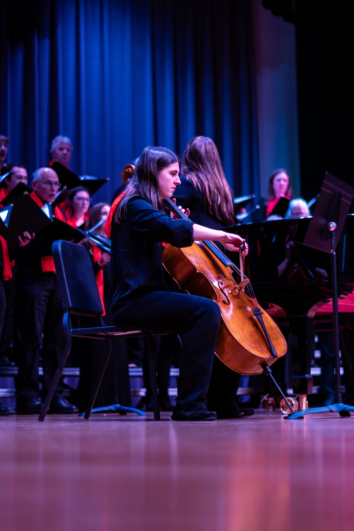 Cellist performs on stage with choir and orchestra members in background under blue stage lighting.