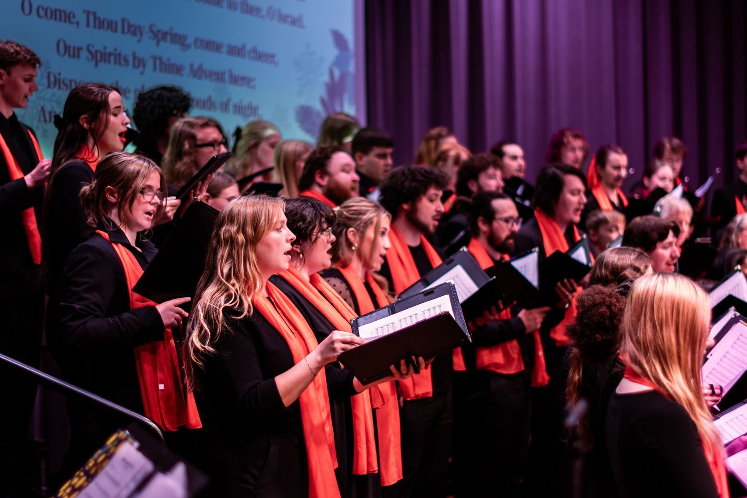 A large choir performs together on stage under dramatic red and purple lighting during a concert performance.