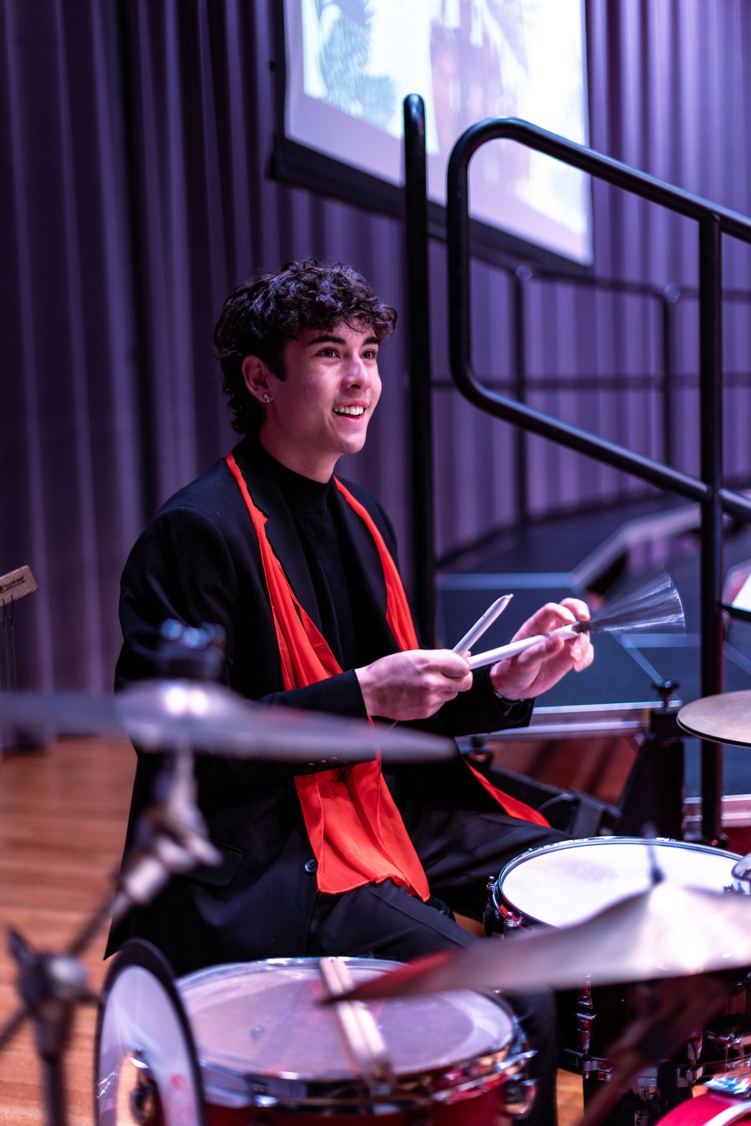 Drummer in black jacket and red scarf performs energetically on stage with drum kit.