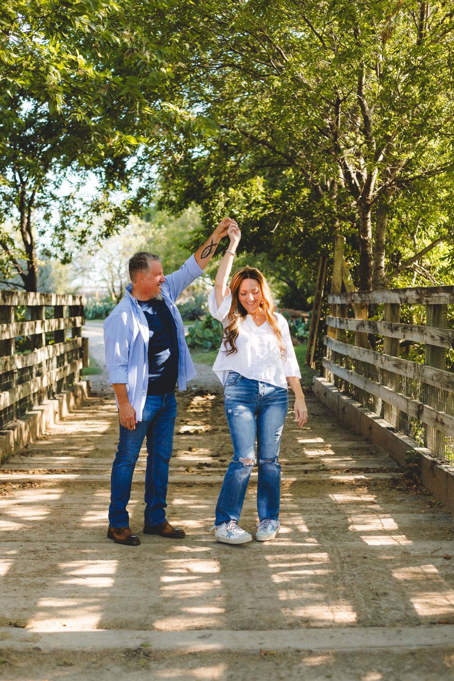 A couple dancing together on a wooden bridge surrounded by lush green trees on a sunny afternoon.