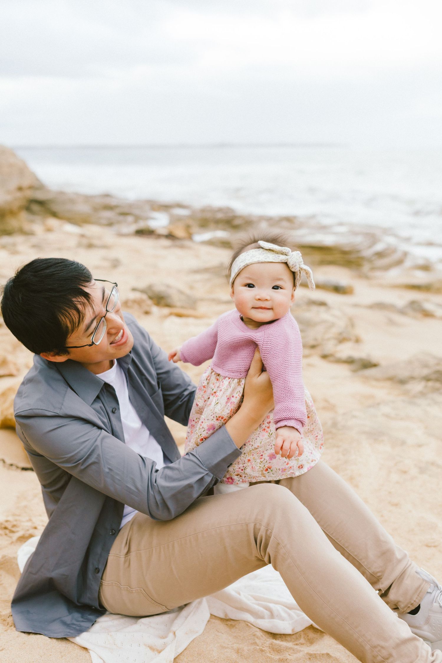 A parent and baby sit together on a rocky beach during sunset, with the baby wearing a pink sweater and headband.