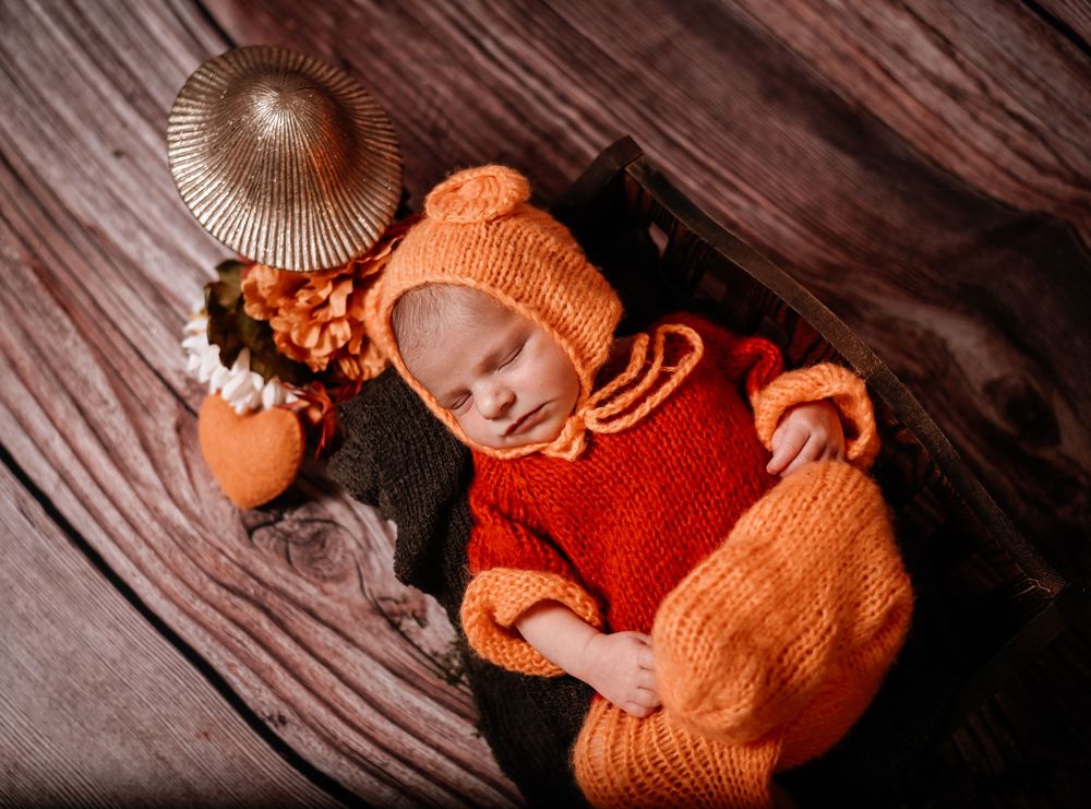 A sleeping baby wearing cozy orange knit clothing and hat lies on a dark wooden surface.