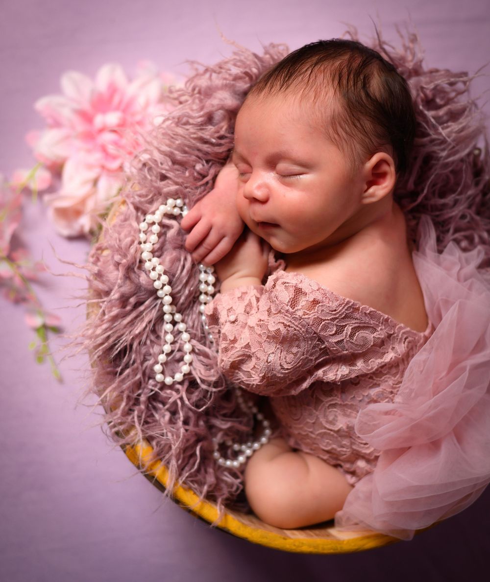 Newborn baby sleeping peacefully on pink tulle fabric with delicate lace accents in soft pink lighting.