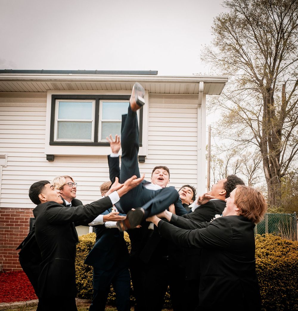 Groomsmen in black suits perform a synchronized jumping pose for a fun wedding photo outdoors.