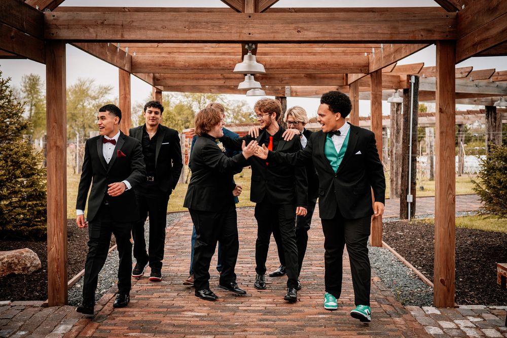 Groomsmen in black suits and teal accents pose playfully under a rustic wooden pergola at an outdoor venue.