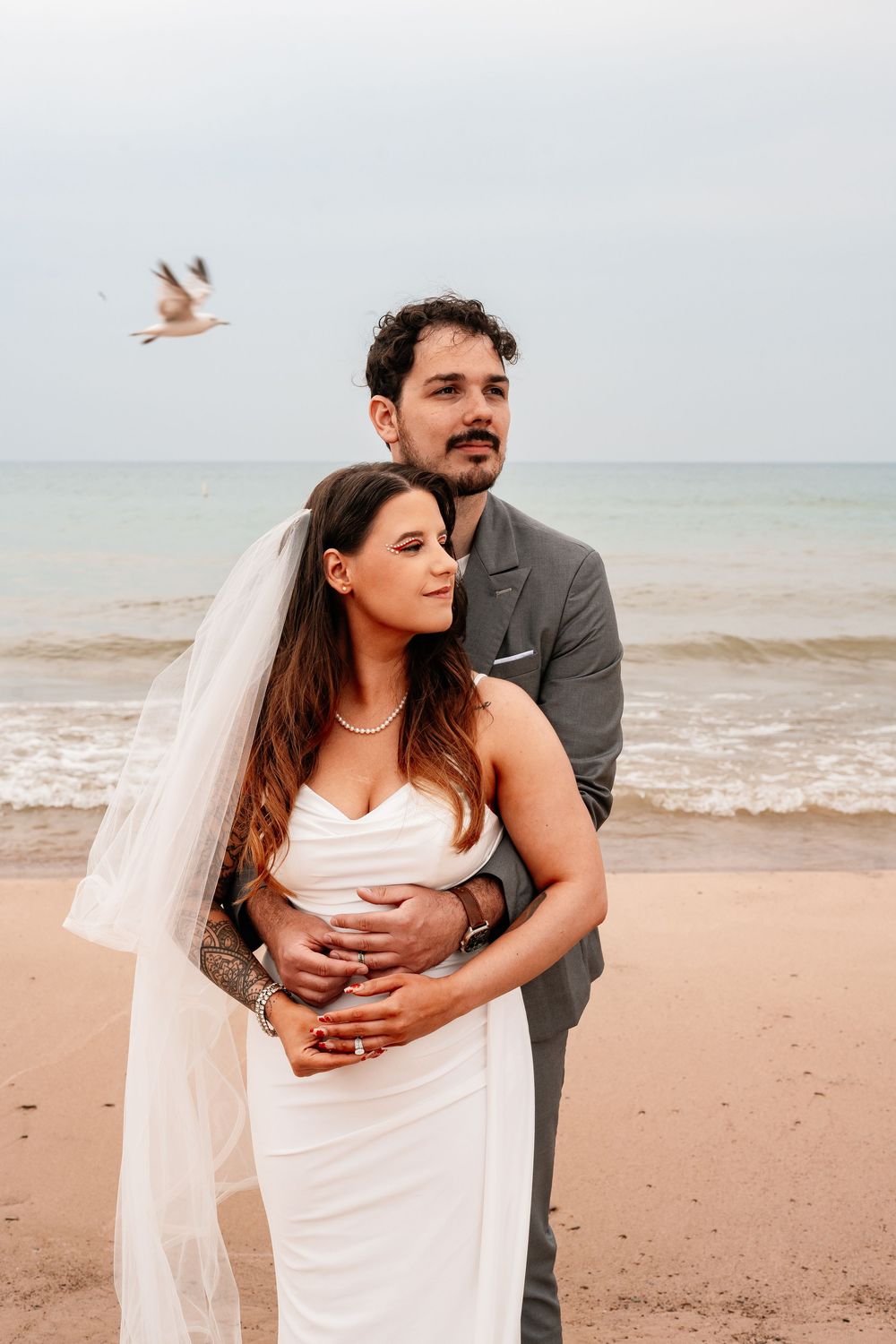 Newlywed couple embraces on a sandy beach with ocean waves in the background.