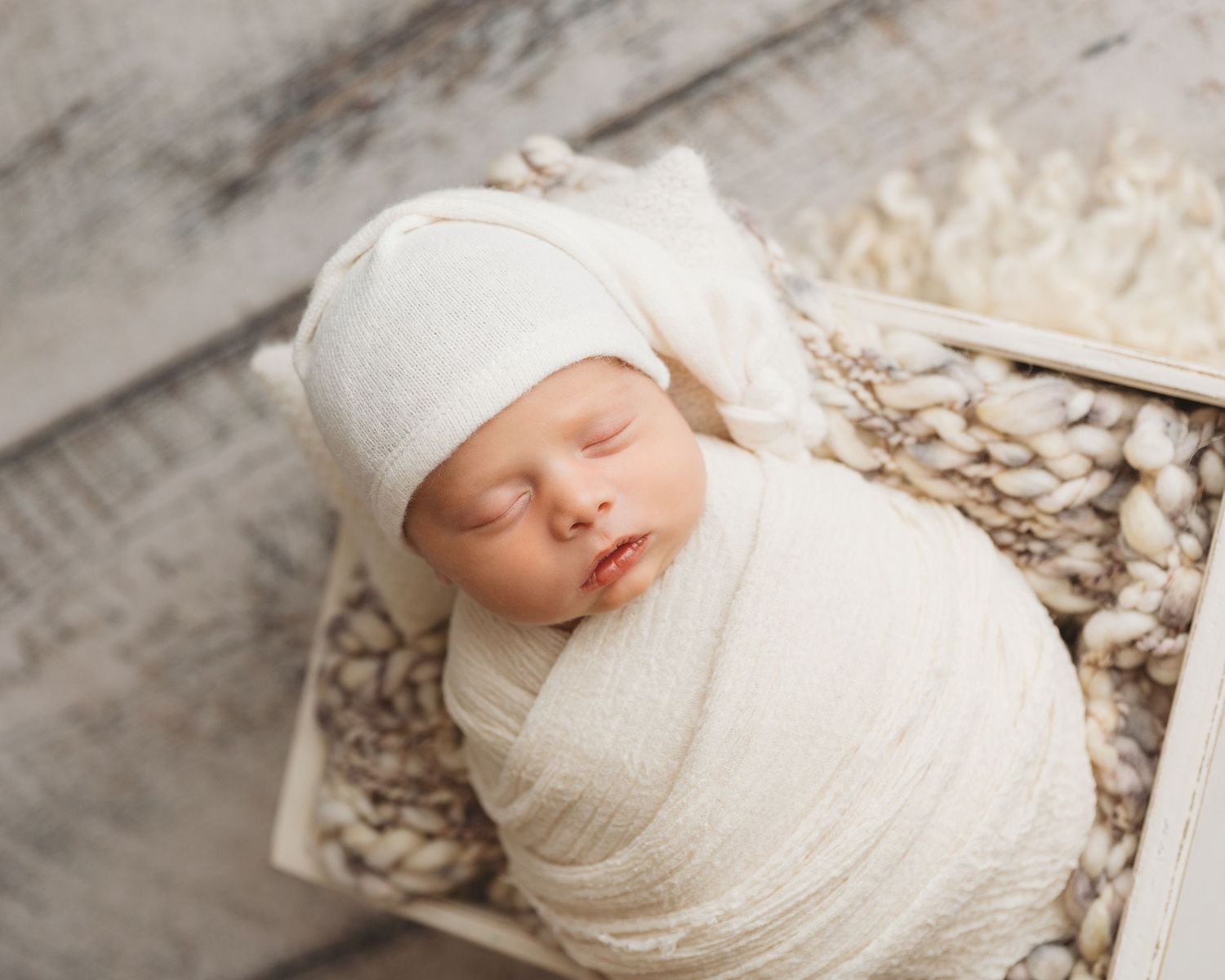 A sleeping newborn baby swaddled in soft white fabric lies on a textured beige backdrop.