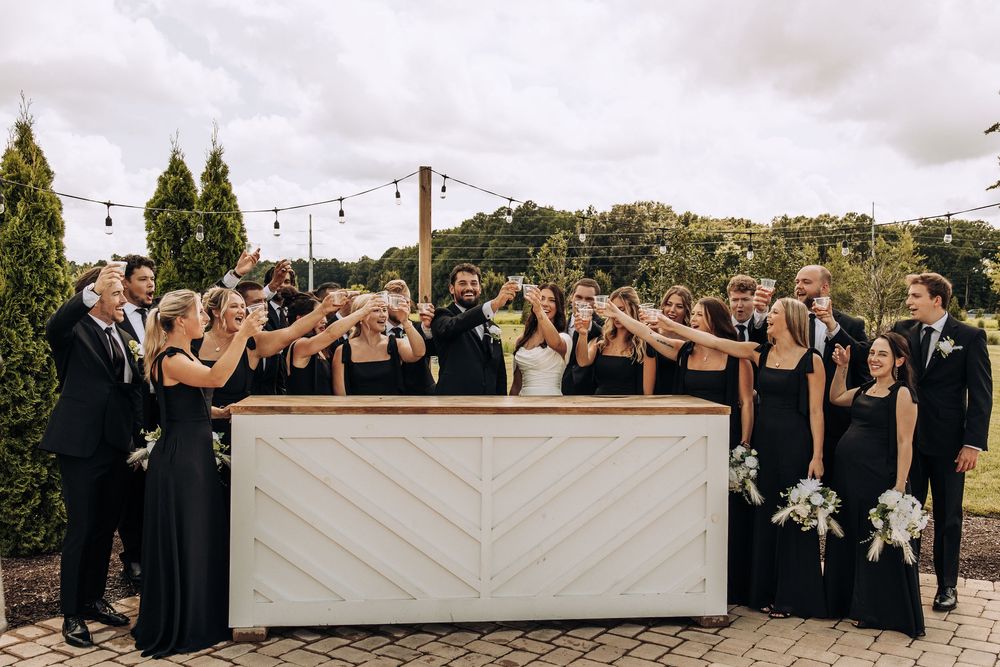Wedding party in black attire toasting drinks at a white chevron bar during outdoor celebration.