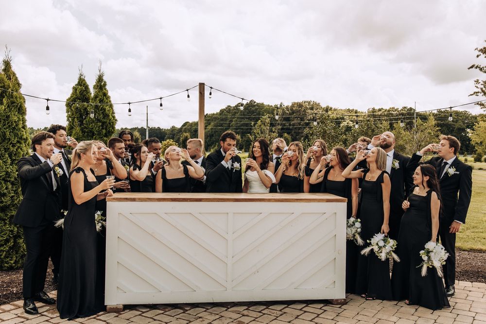 Large wedding party in black formal wear toasting at white chevron outdoor bar.