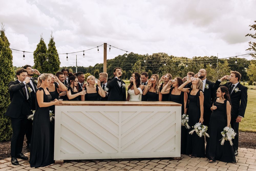 Wedding party laughing and toasting drinks together at white outdoor bar during reception.