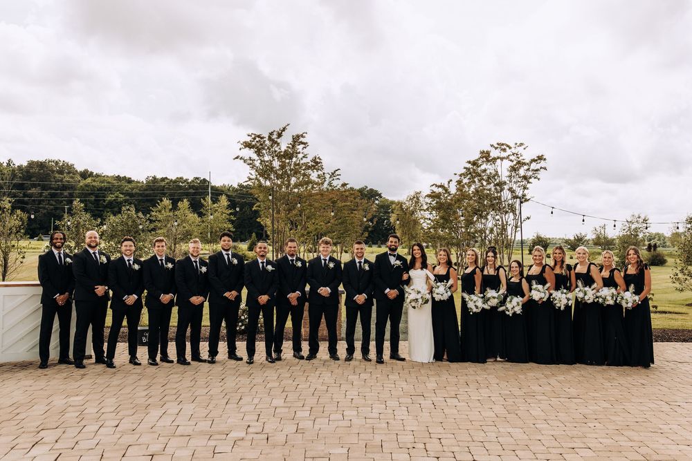 Large wedding party in black formal attire posing outdoors on brick patio with greenery.