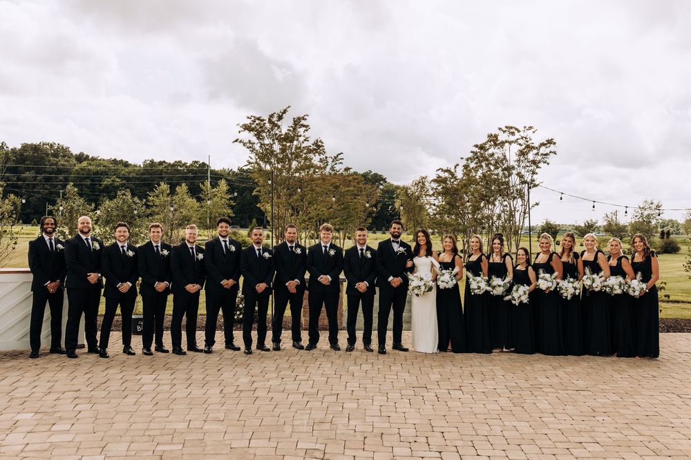 Bride and groom pose with large wedding party in matching black attire on outdoor patio.