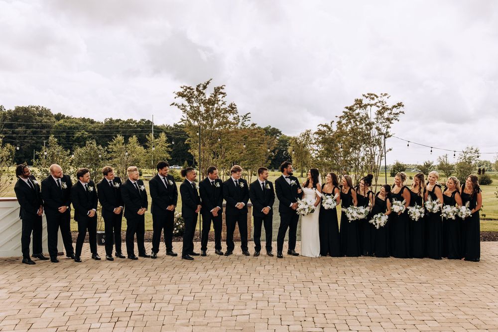 Elegant wedding party in all black formal wear posing on brick patio with lush greenery.