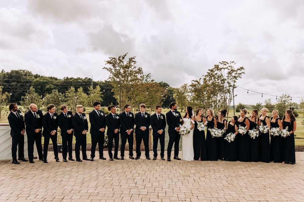 Large wedding party in black formal attire posing on brick patio under cloudy sky.