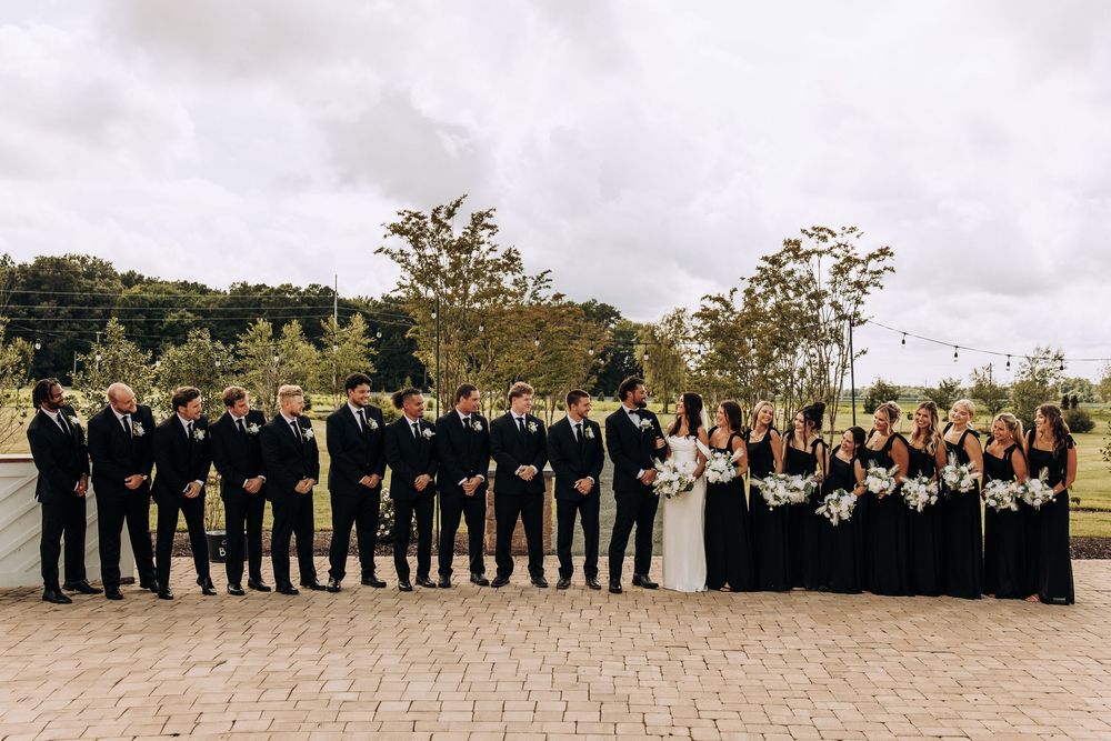 Large wedding party in black attire with bride and groom centered on brick patio with green landscape backdrop.