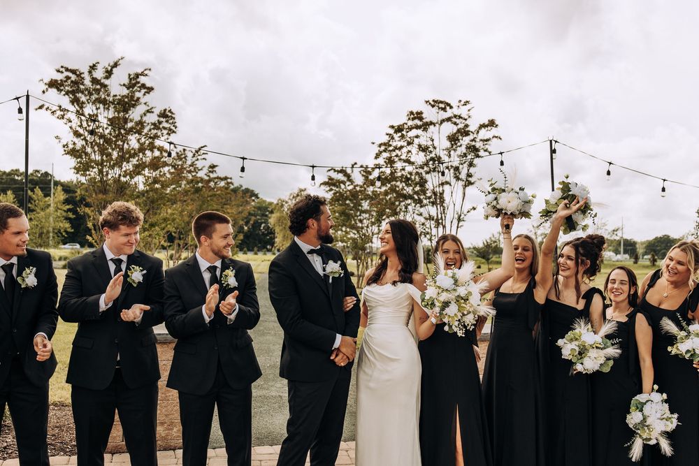 Wedding party in black laughing and celebrating with bride and groom outdoors under string lights.