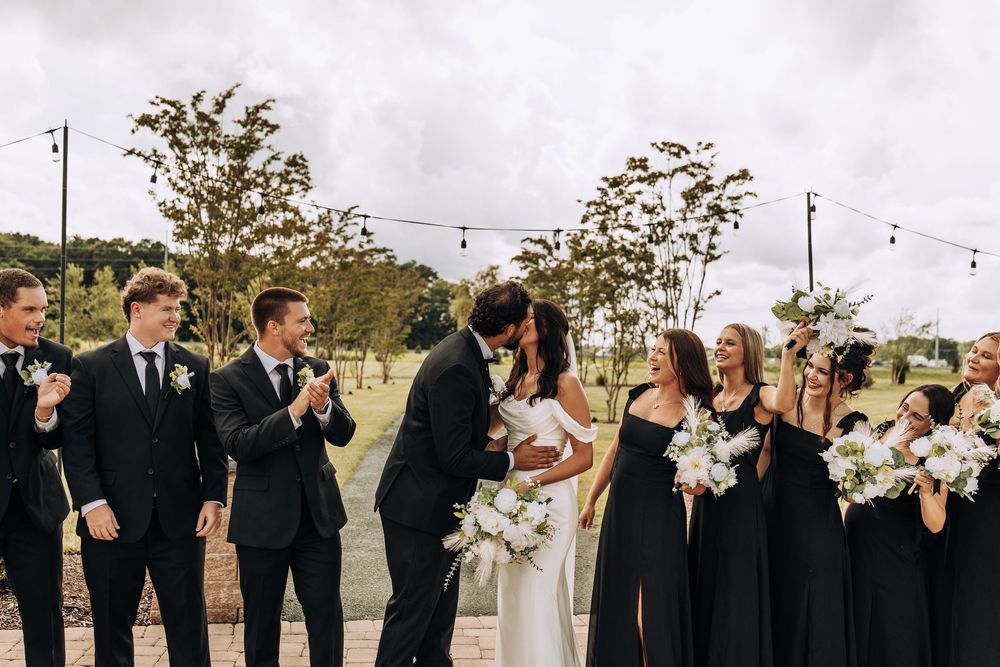 Bride and groom share a kiss surrounded by cheering wedding party in black formal attire outdoors.