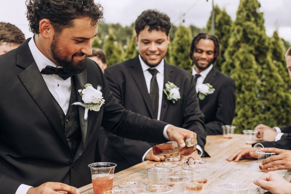 Groomsmen in black suits pour drinks at outdoor wedding reception table with white boutonnieres.