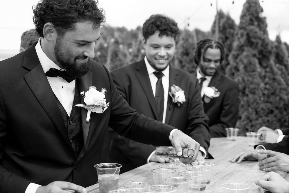 Black and white photo of groomsmen laughing and pouring drinks at outdoor wedding reception table.