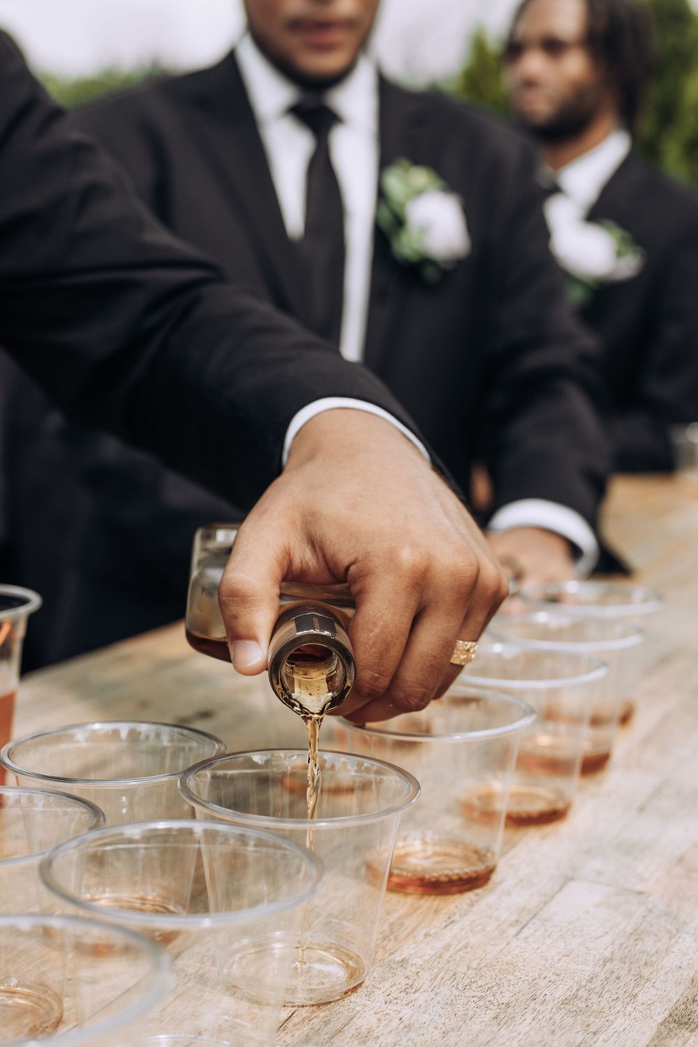 Groomsman pouring whiskey into plastic cups on a wooden table at an outdoor wedding reception.