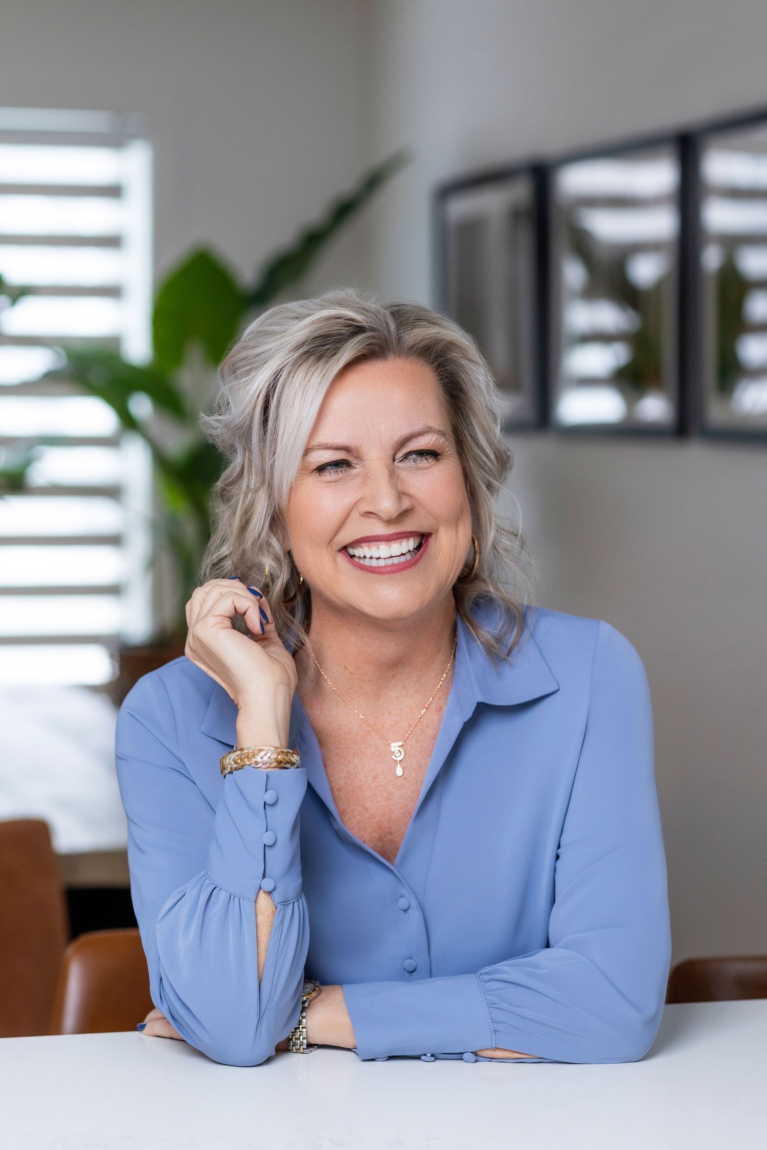 Professional portrait of someone wearing a light blue button-up shirt smiling warmly in a bright modern office setting.