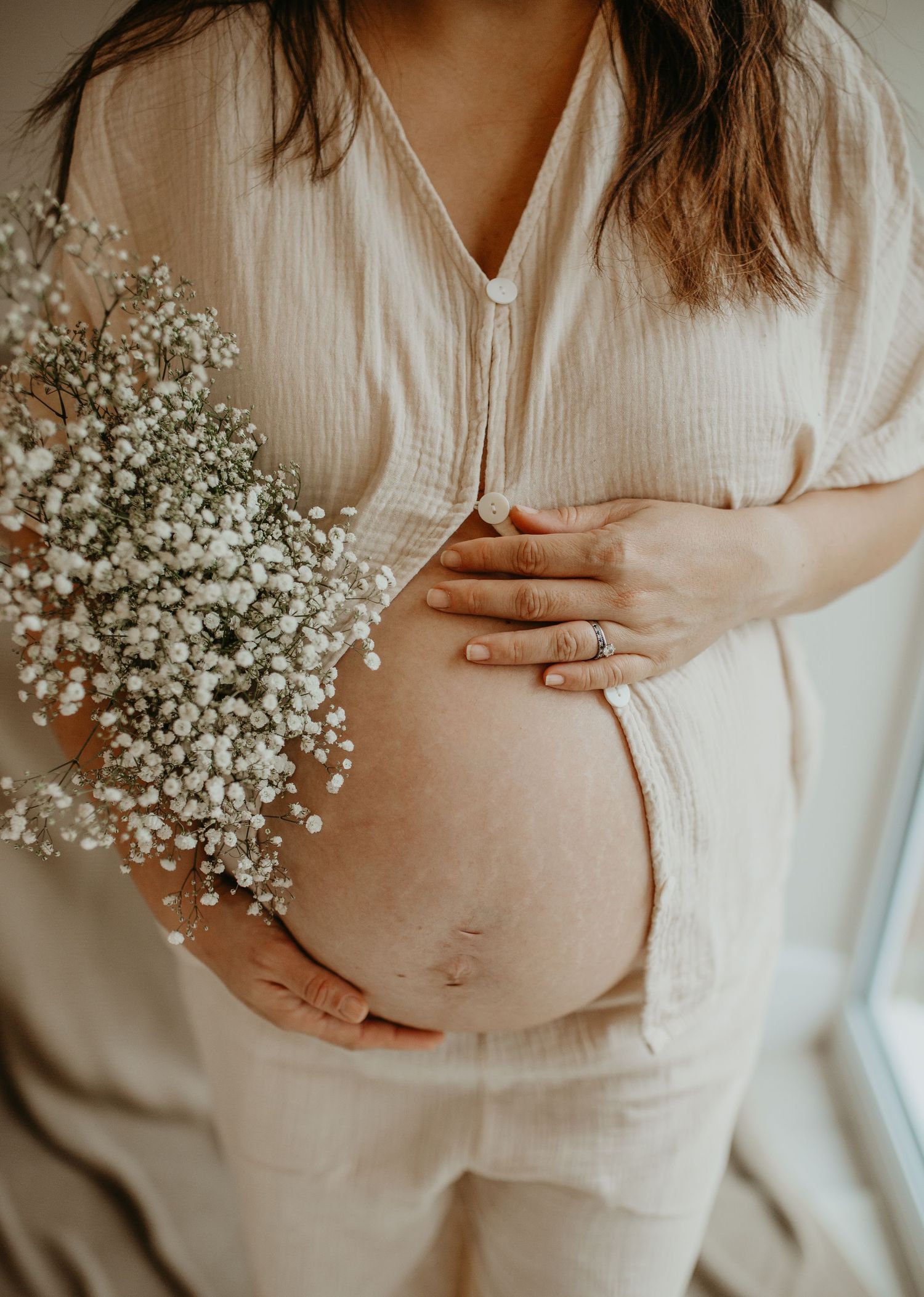 Pregnant woman in beige linen outfit cradling baby bump while holding delicate white baby's breath flowers.