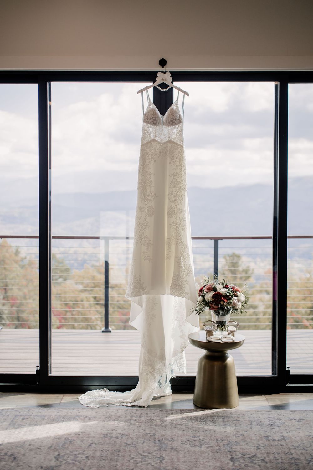 White lace wedding dress hanging in front of large windows with mountain view.