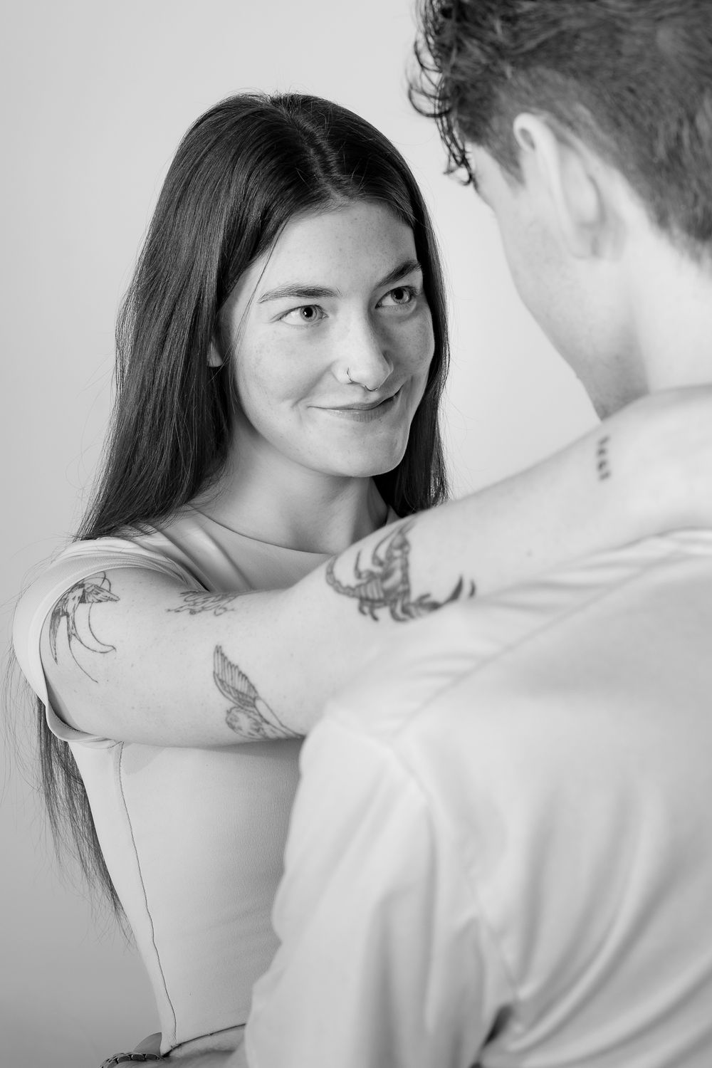 Black and white portrait of a couple with visible tattoos sharing an intimate moment against a plain background.