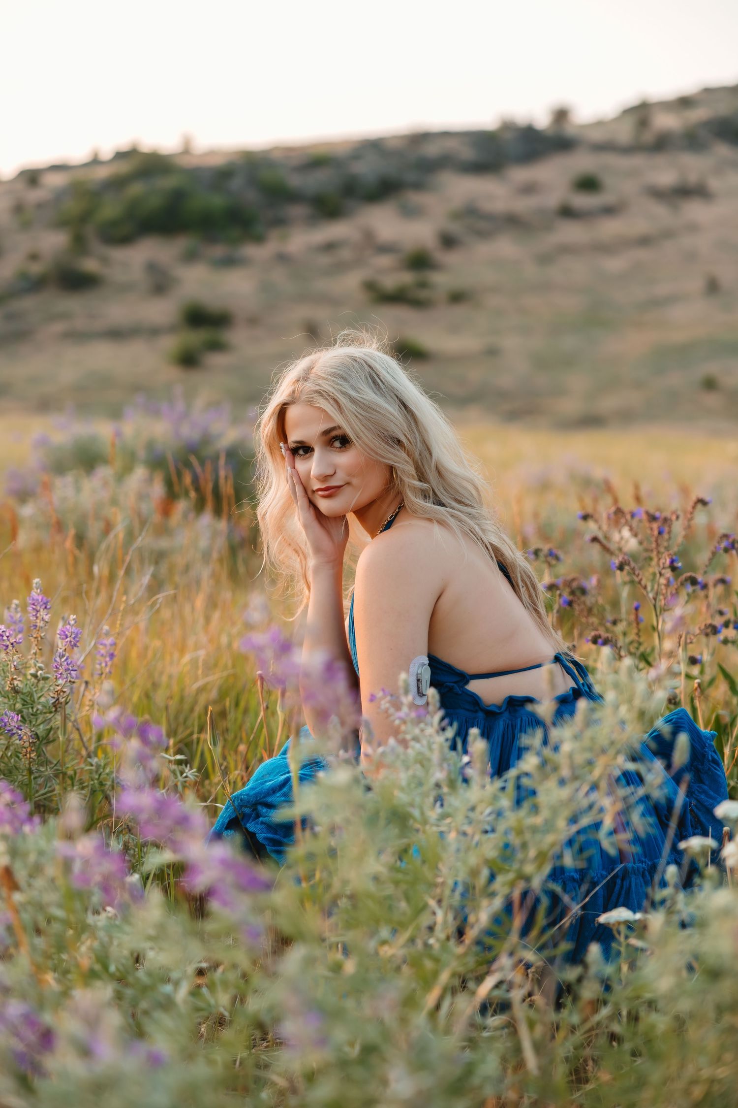 A person in a blue dress poses gracefully among purple wildflowers in a desert landscape during golden hour.
