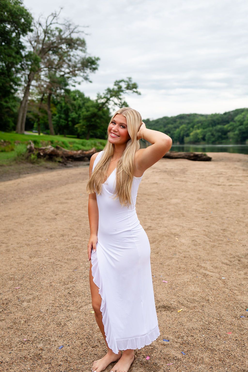 Person in a flowing white dress poses on a sandy beach shore near a lake surrounded by green trees and hills.