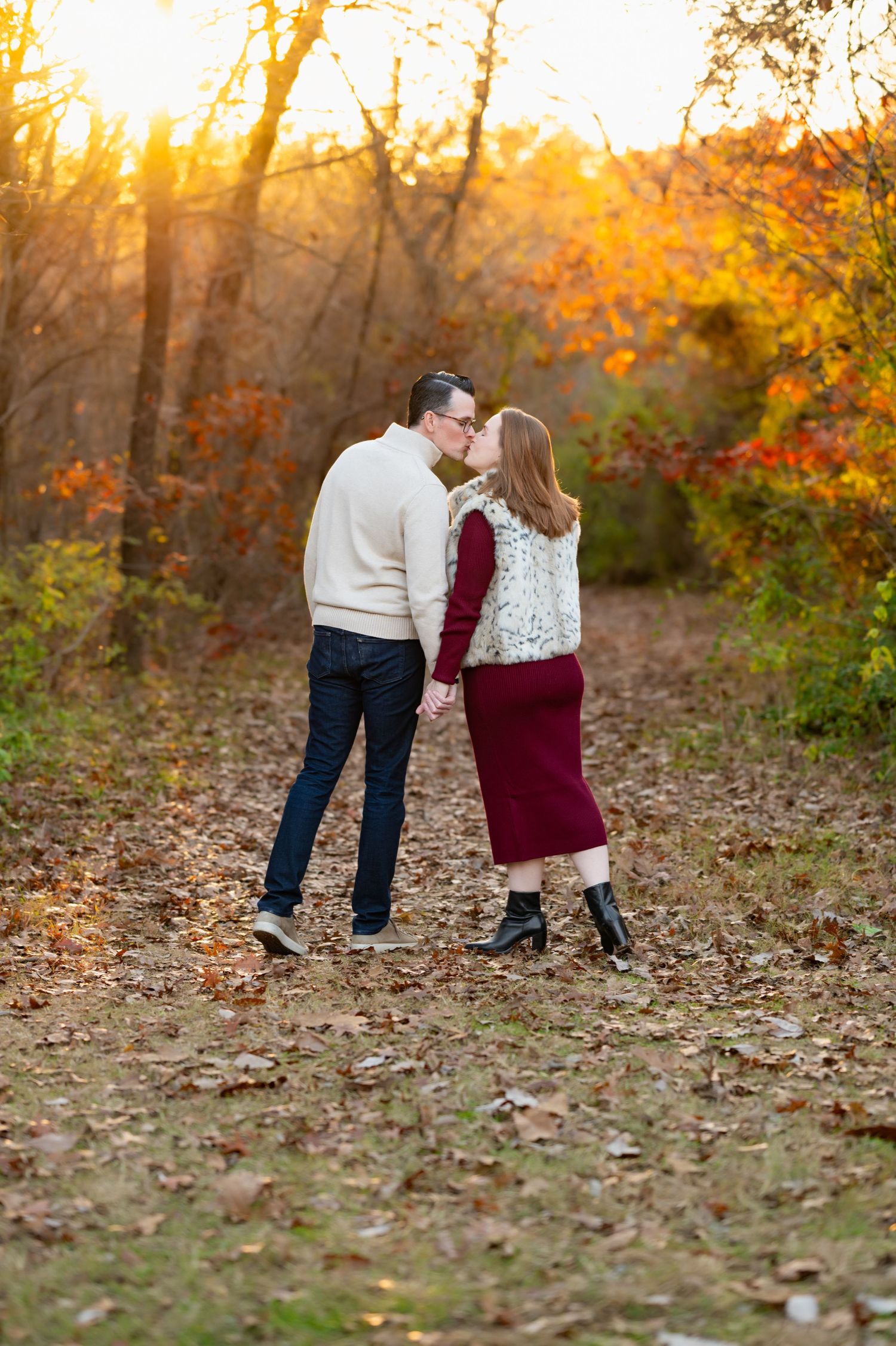 soon-to-be parents holding hands walking away from camera into the sunlight of a wooded area pause for a peck during dallas maternity photos