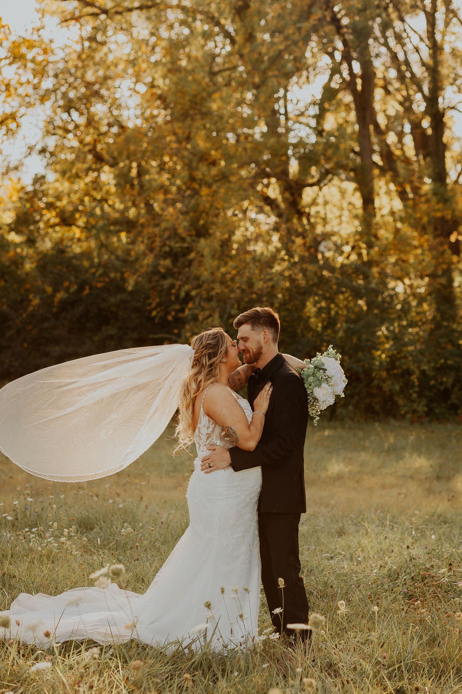A romantic sunset wedding photo with a flowing veil catching the golden light as the couple embraces in a meadow.