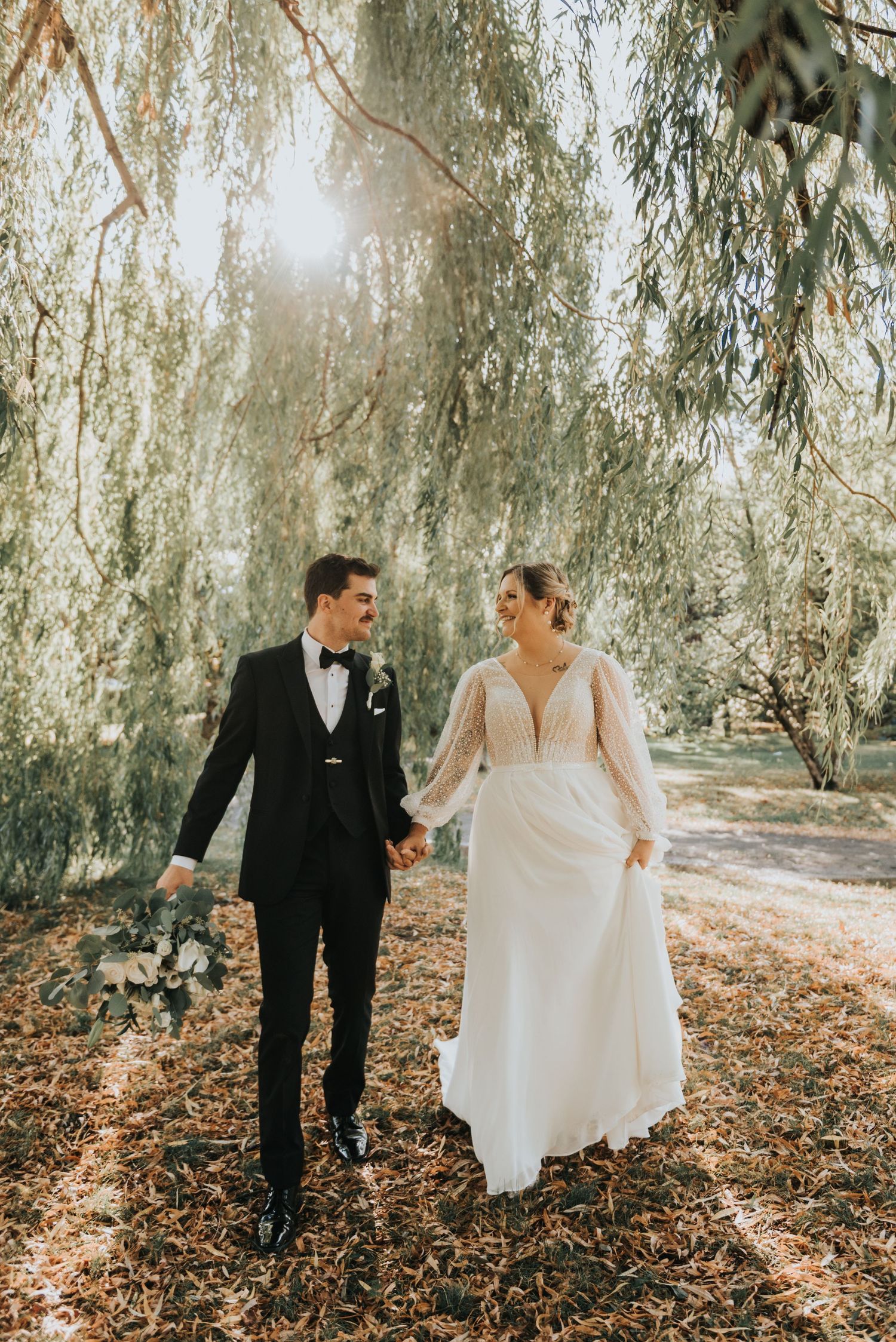 Newlyweds walk together under cascading willow trees on a sunlit autumn day.