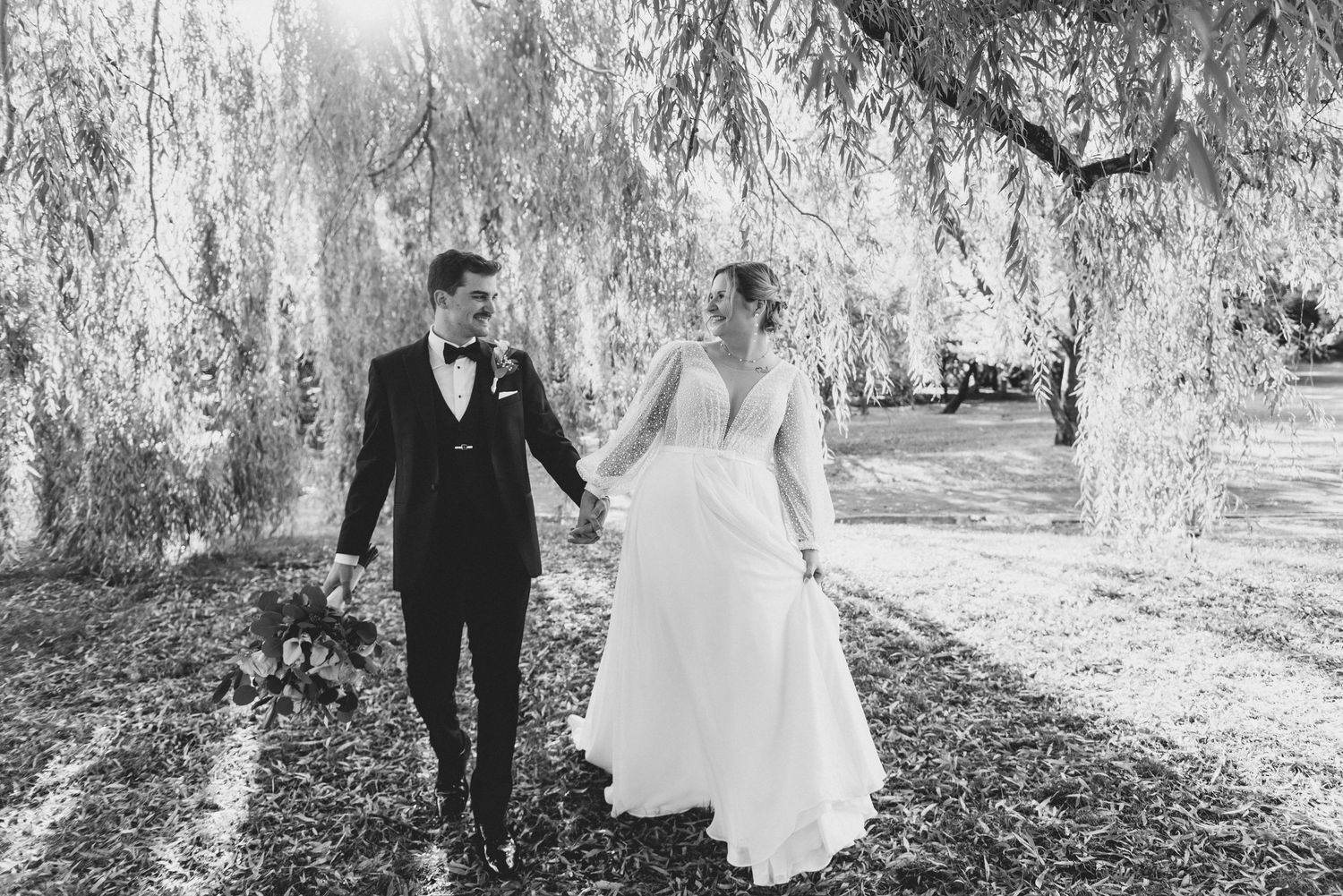 Black and white wedding photos show a couple walking together under weeping willow trees in a romantic outdoor setting.