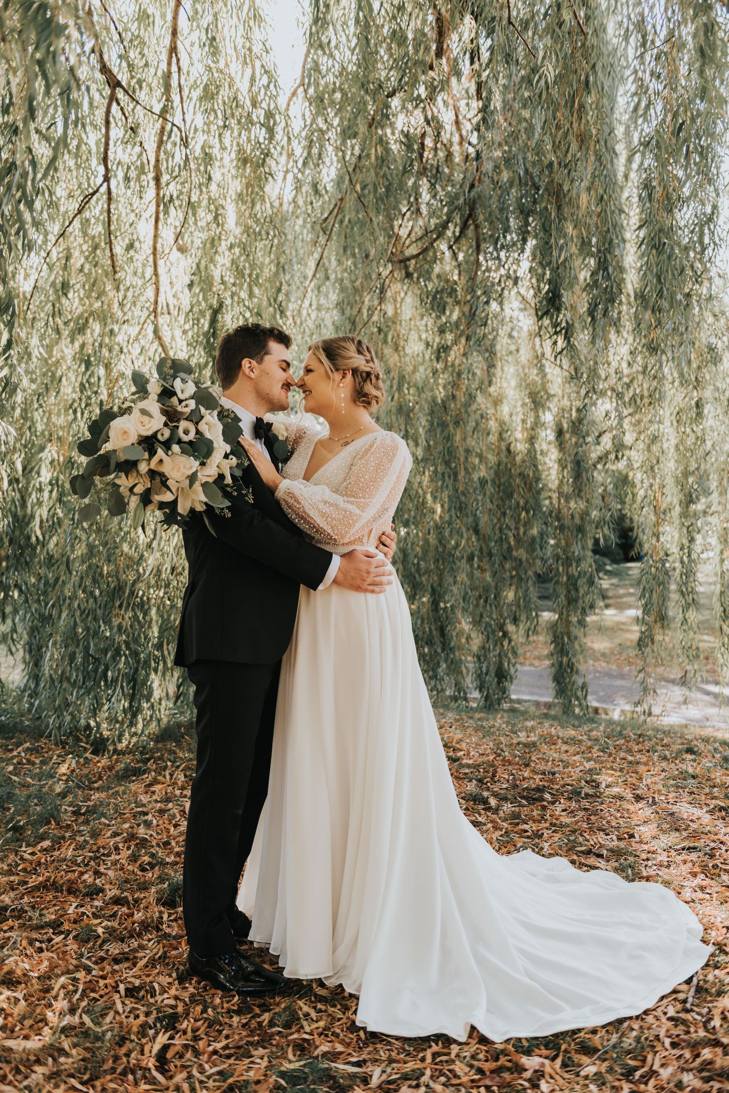 A romantic wedding moment under a willow tree with flowing white dress and autumn leaves on the ground.