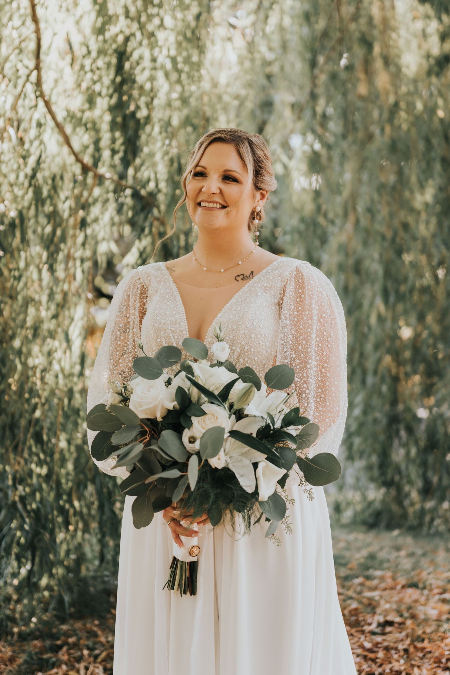 A bride in a flowing white lace dress holds a beautiful bouquet of white roses and eucalyptus leaves in a wooded setting.