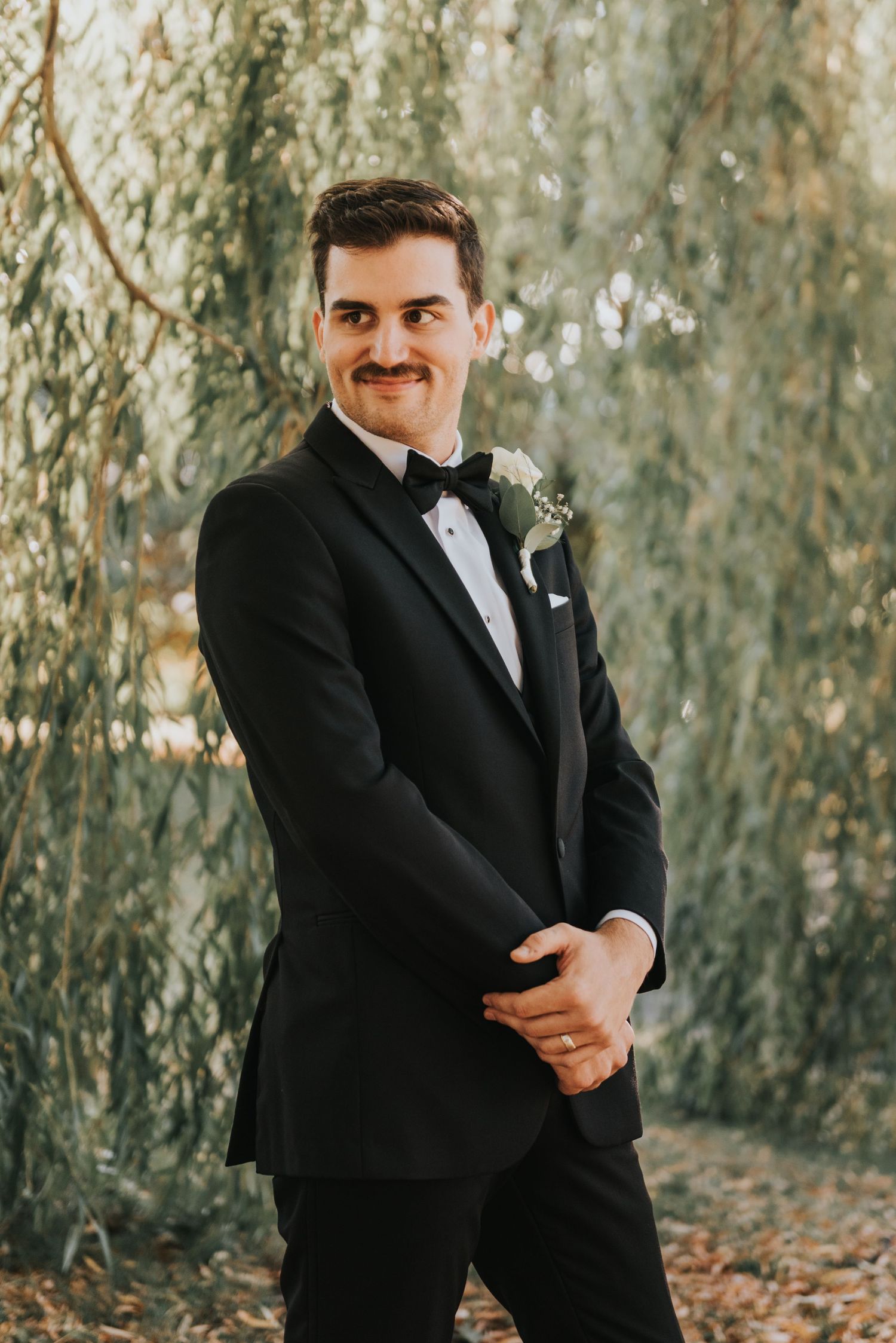 A groom in a classic black tuxedo poses among willow trees during an autumn wedding photoshoot.
