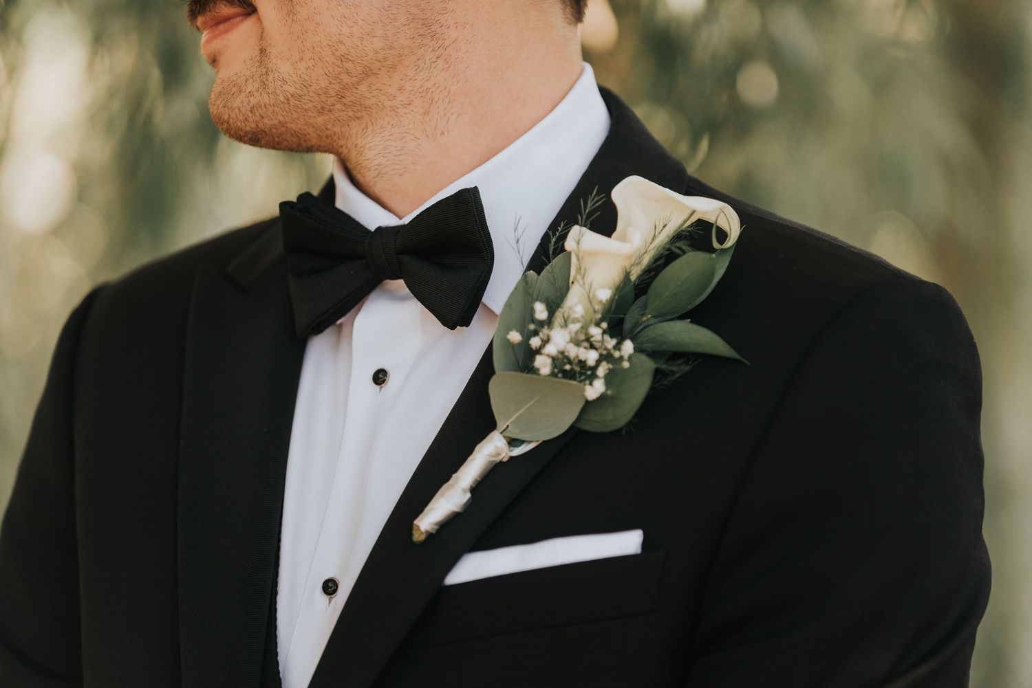 Groom in black tuxedo and bowtie adjusts his boutonniere during wedding preparations.