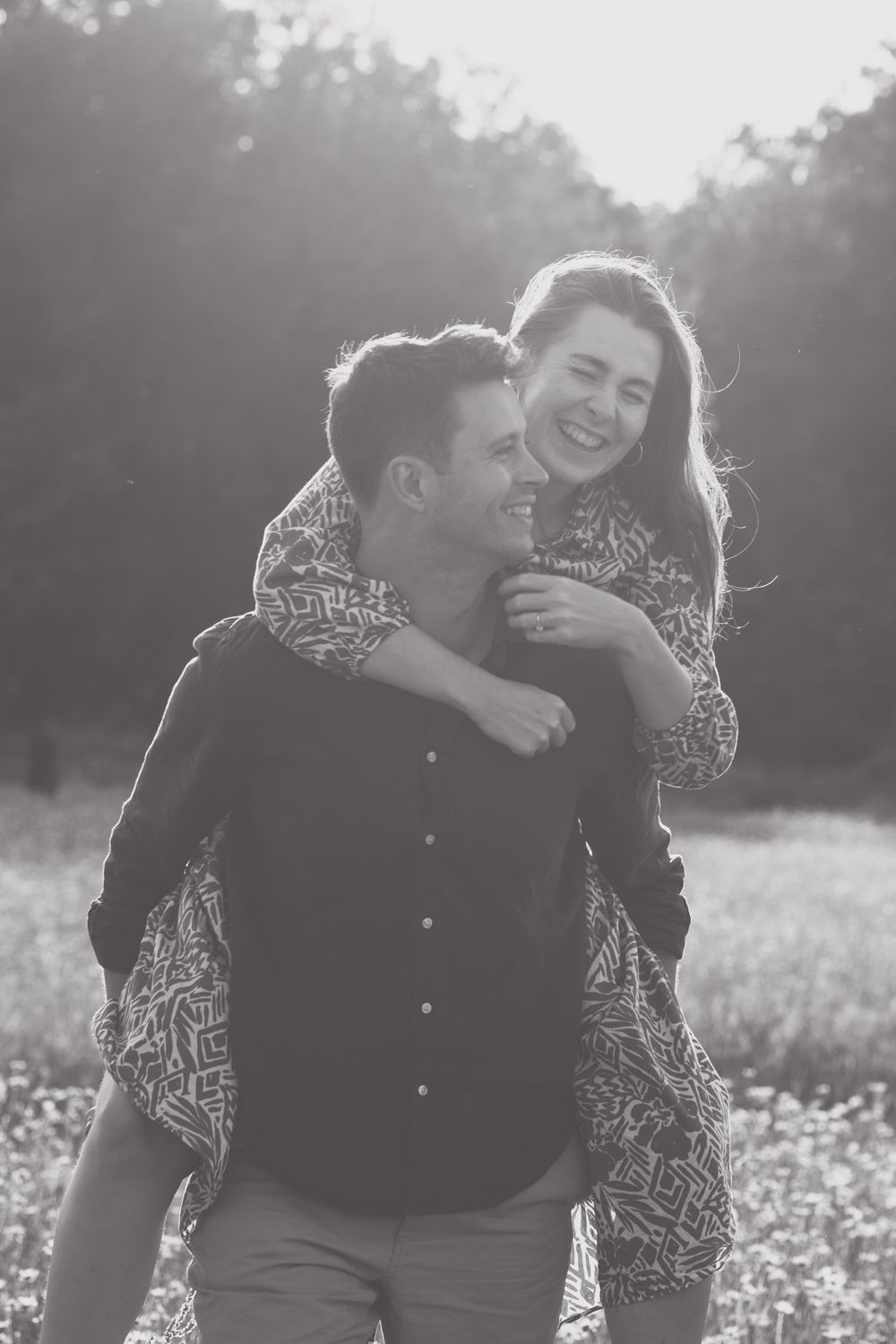 A couple shares joyful laughter together in a sunny field during a black and white photoshoot.