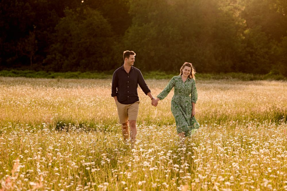 A couple walks hand in hand through a golden meadow filled with wildflowers during sunset.