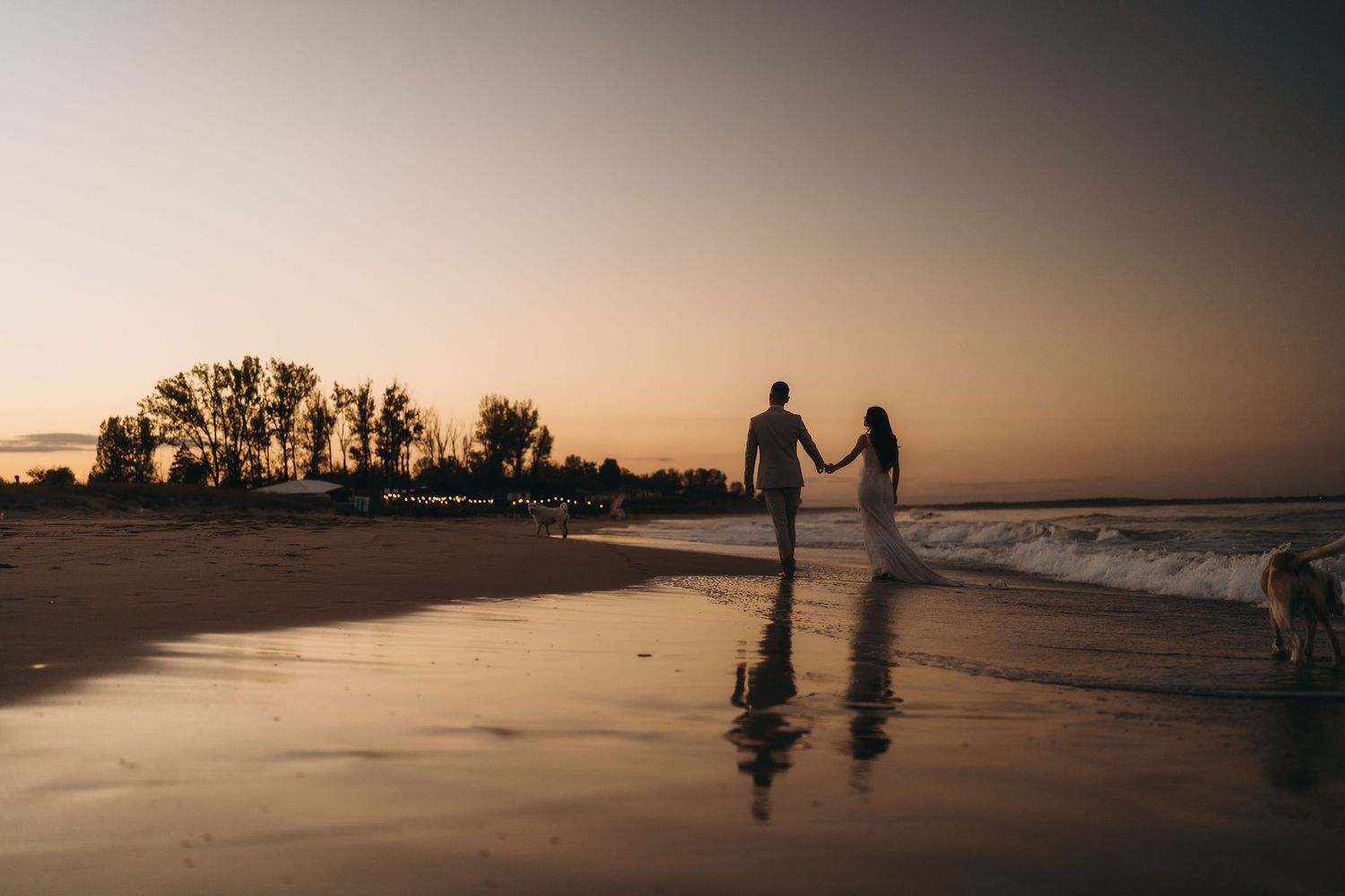 Silhouettes of couple holding hands on beach at sunset with reflections in wet sand.