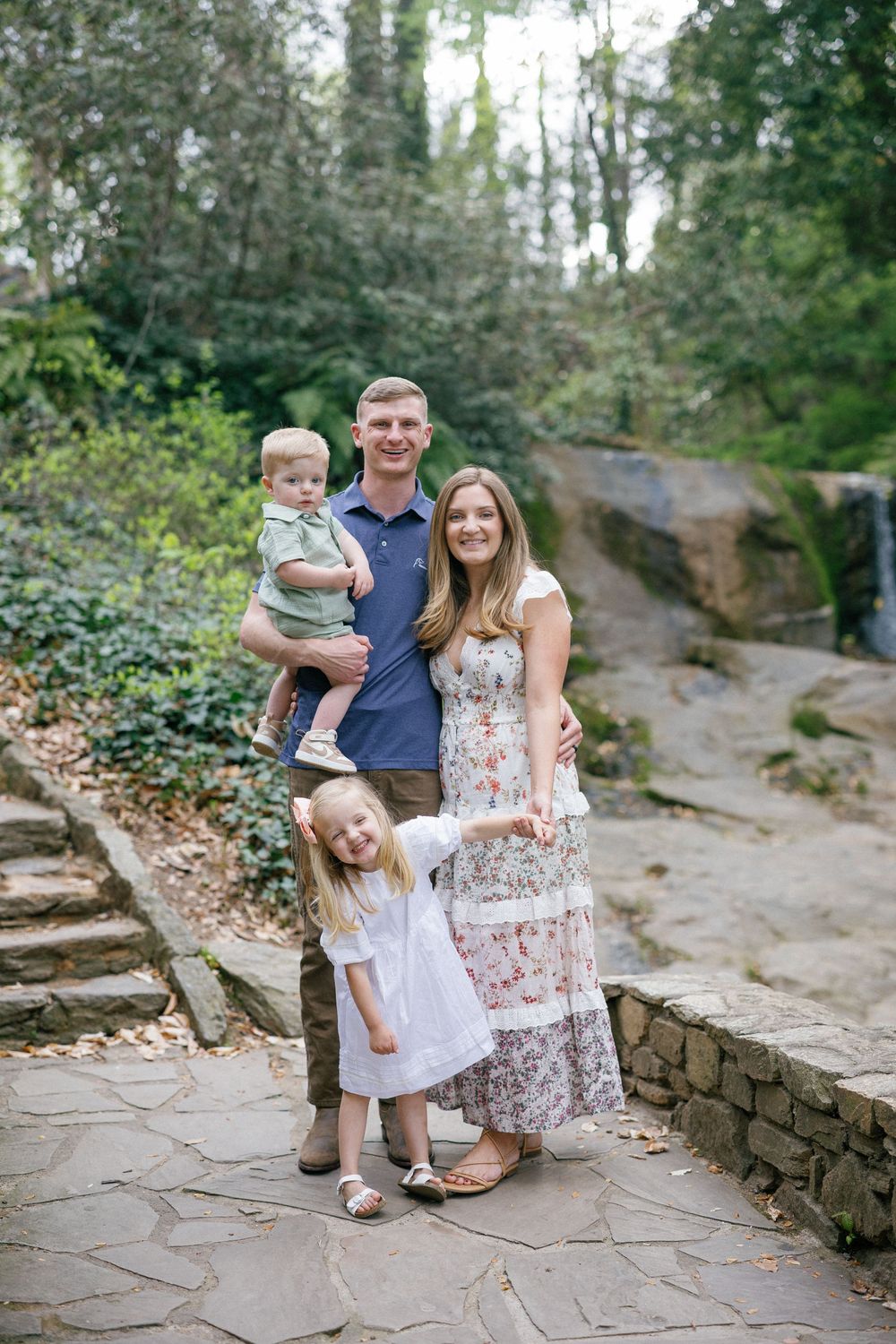 A happy family of four poses on a stone path surrounded by lush greenery and a waterfall backdrop.