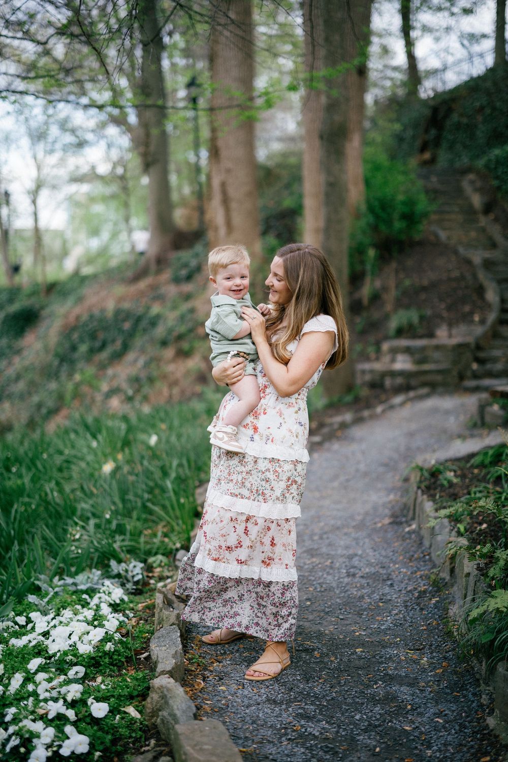 A young mother in a floral maxi dress holds her baby boy on a garden path surrounded by lush greenery.
