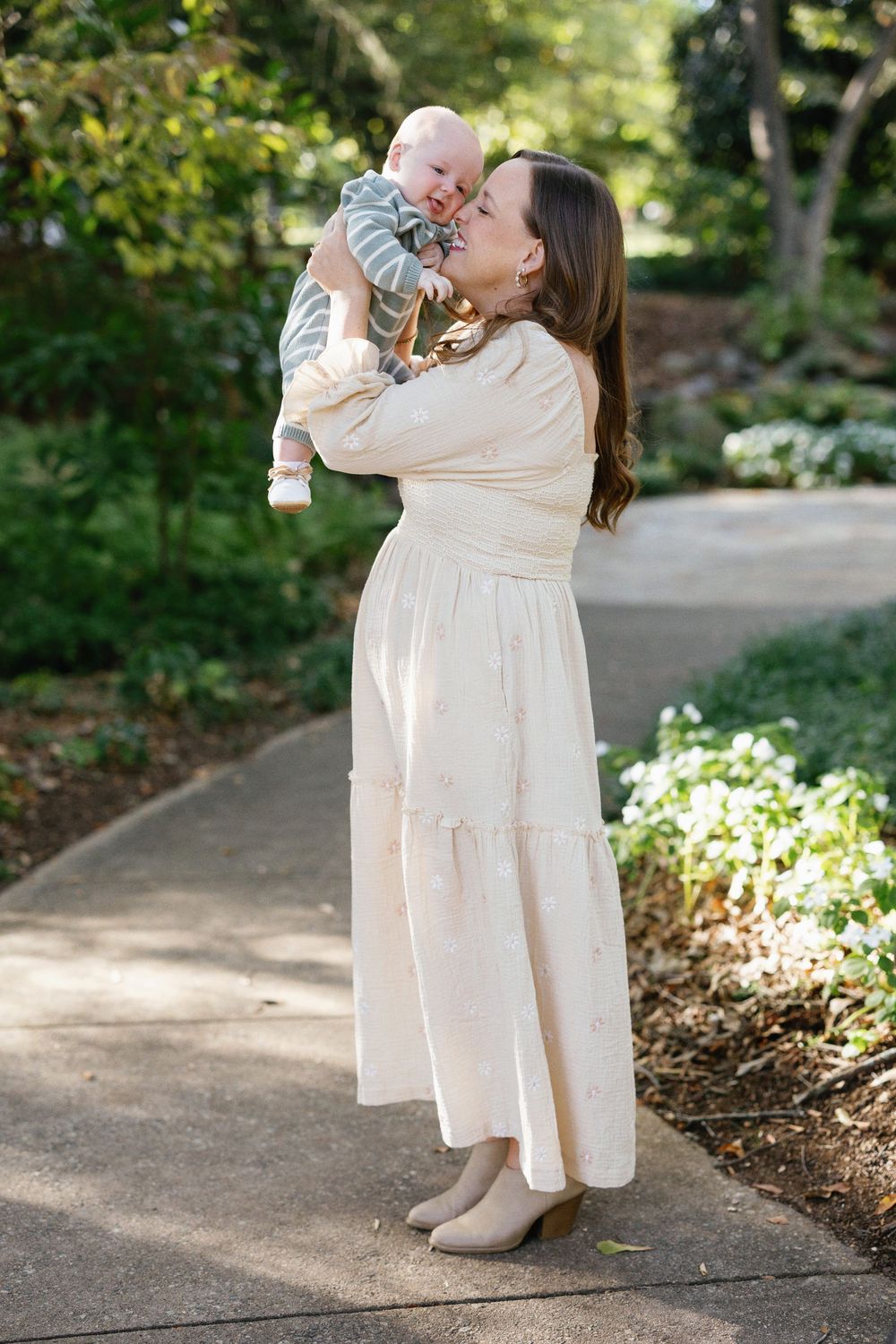 A parent in a white dress holds and lifts a small baby outdoors in a garden setting during a sunny day.