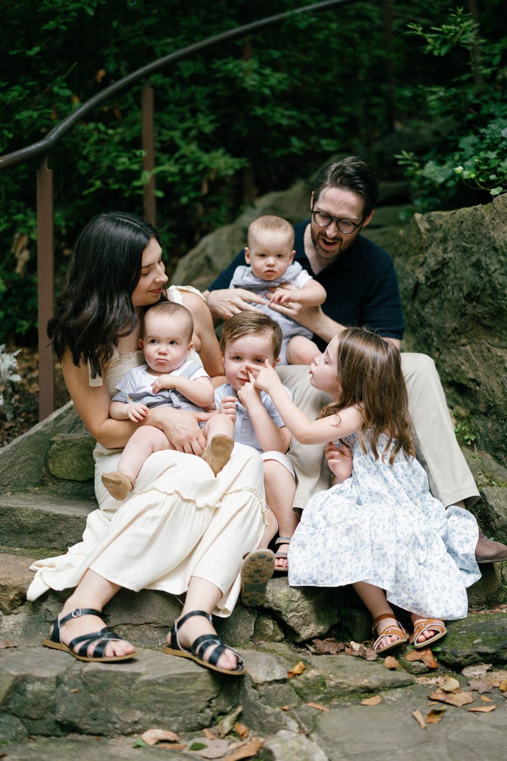 Family sitting together on stone steps in a wooded area wearing coordinated white and cream colored outfits.