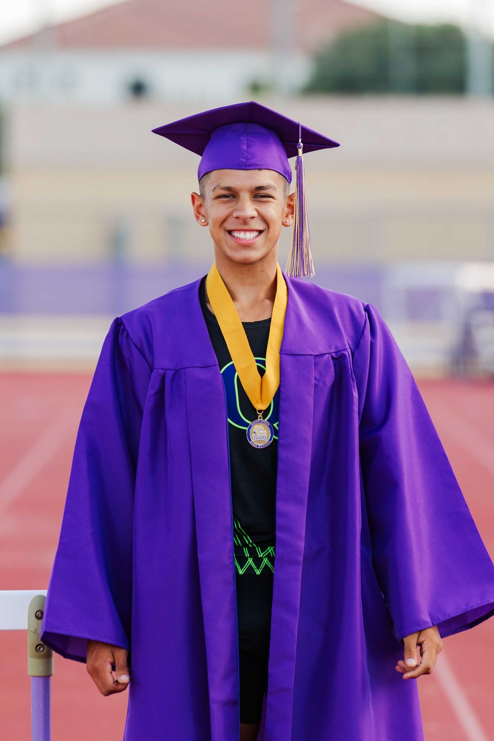 Graduate wearing purple cap and gown with yellow stole stands proudly at outdoor graduation ceremony.