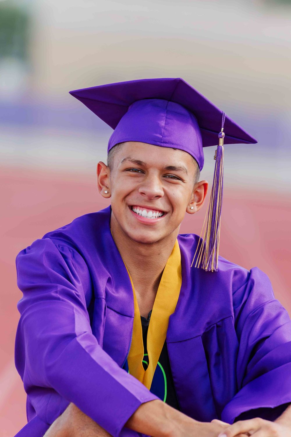 Graduate wearing purple cap and gown with gold stole sits on track field during graduation photo session.