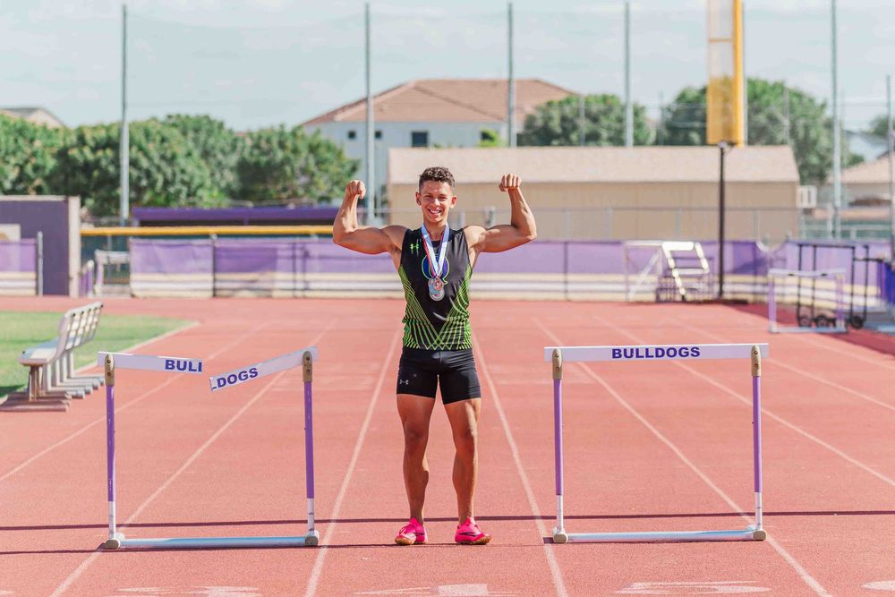 Track athlete stands victoriously between hurdles on running track wearing medals and athletic gear.