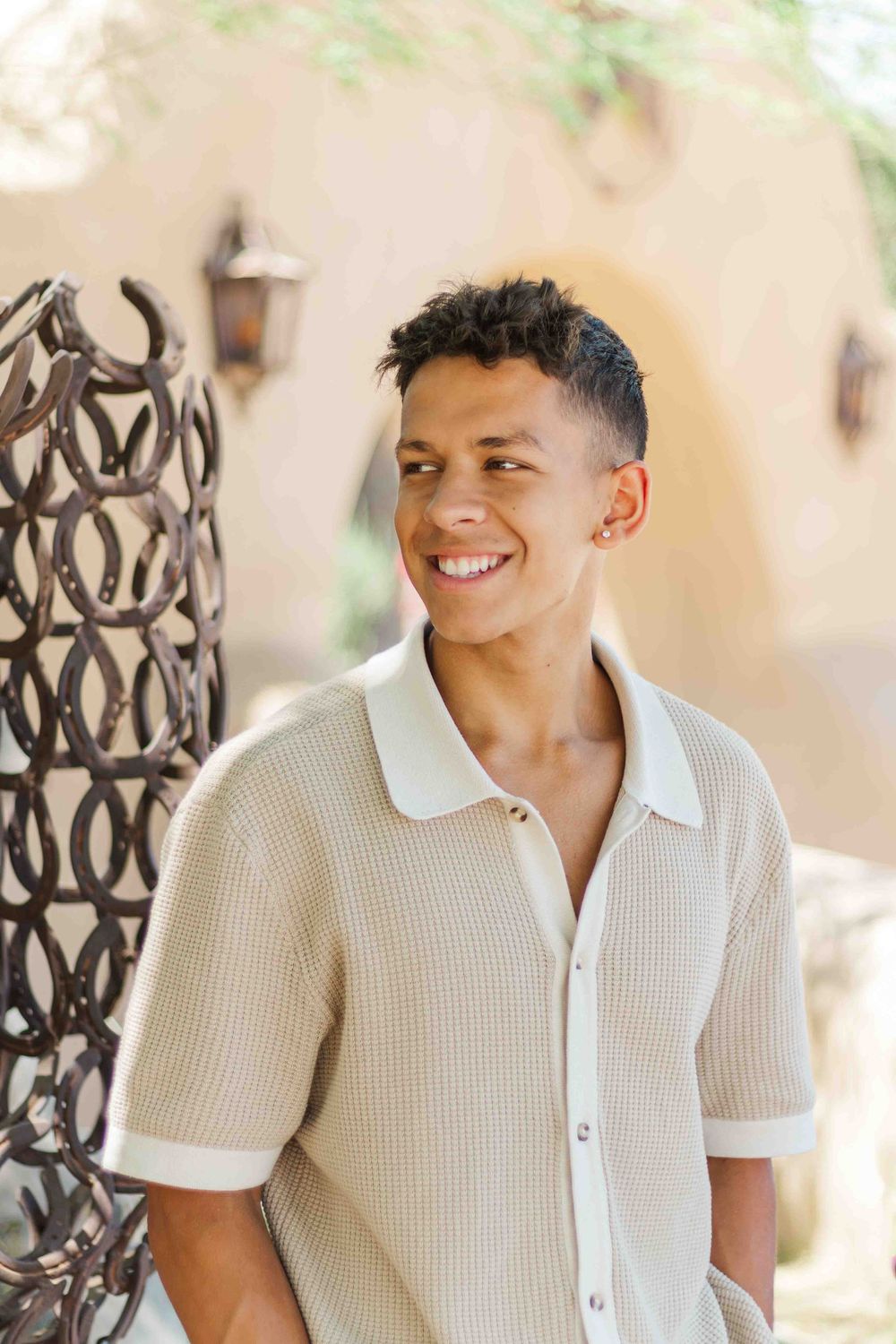 A smiling person wearing a cream-colored knit polo shirt stands against a blurred outdoor background with ornate ironwork.