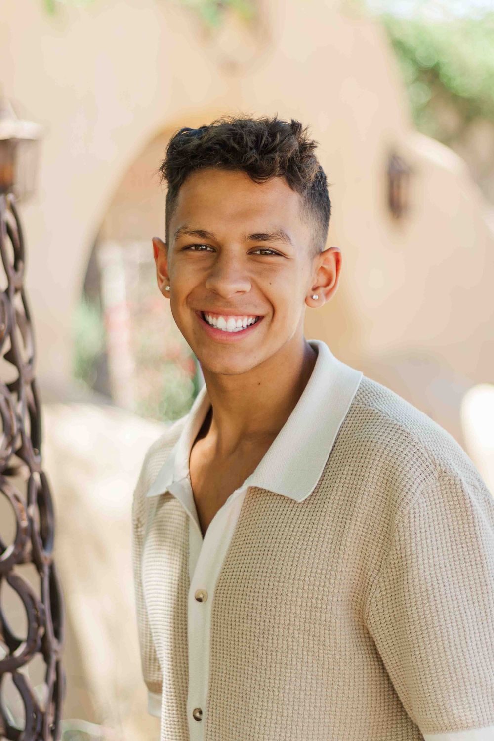 A person wearing a light colored textured shirt smiles warmly in an outdoor setting with soft natural lighting.